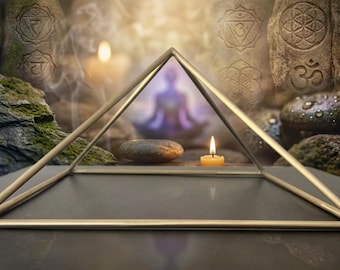 May include: A gold-toned metal pyramid frame with a square base. The structure consists of four thin, straight metal rods meeting at a single apex point, creating a hollow geometric shape that sits on a flat, dark, reflective surface.