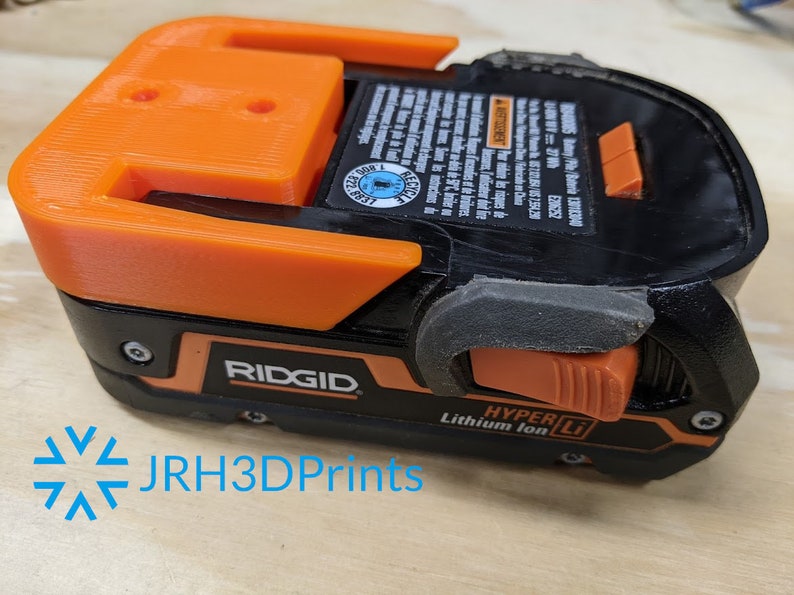 K&ouml;nnte beinhalten: Ein orange-schwarzer Ridgid-Akku mit dem Text "HYPER Li Lithium Ion" an der Seite. Der Akku hat einen schwarzen Kunststoffgriff und einen schwarzen Kunststoffverschluss.
