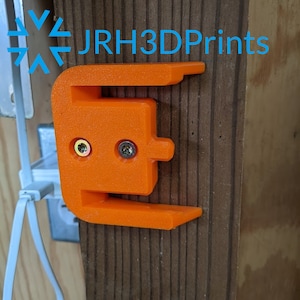 K&ouml;nnte beinhalten: Orangefarbener 3D-gedruckter Wandhalter mit zwei Schraubenl&ouml;chern. Der Halter ist an einer Holzoberfl&auml;che befestigt. Der Text "JRH3DPrints" ist im Hintergrund sichtbar.