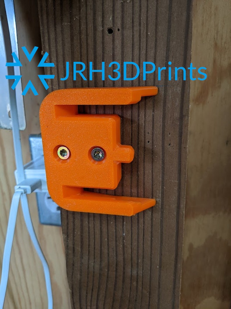 K&ouml;nnte beinhalten: Orangefarbener 3D-gedruckter Wandhalter mit zwei Schraubenl&ouml;chern. Der Halter ist an einer Holzoberfl&auml;che befestigt. Der Text "JRH3DPrints" ist im Hintergrund sichtbar.