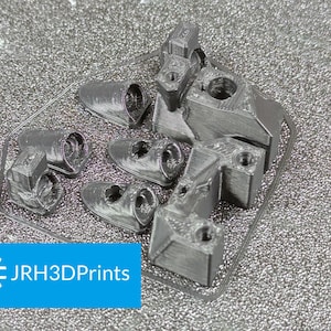 May include: A collection of gray 3D-printed components, including cylindrical and rectangular shapes with holes and textured surfaces. The components are arranged on a textured, silver surface. The image includes a blue rectangle with the text "JRH3DPrints".