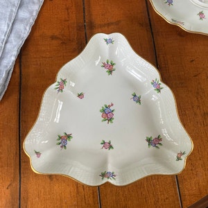 Puede incluir: Una fuente de servir blanca, triangular, con borde festoneado y ribete dorado. El plato está decorado con pequeños racimos de flores rosas, azules y moradas. El plato tiene una textura tejida.
