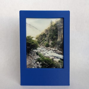 Puede incluir: Un marco de fotos azul con una foto de un río que fluye a través de un bosque. La foto está en orientación horizontal.