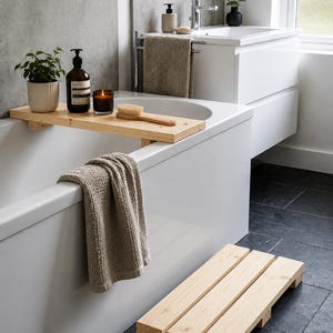 May include: A light-colored wooden bathtub tray resting across a white bathtub, holding a small potted plant, a dark brown pump bottle, a glass candle, and a wooden bath brush. A matching wooden slatted bath mat sits on the dark floor.