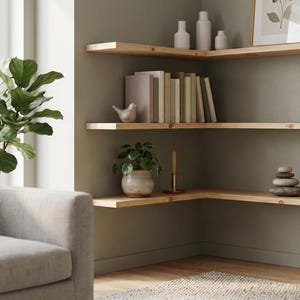 Op de afbeelding: Drie hoekplanken van lichtgekleurd hout gemonteerd aan een saliegroene muur. De planken bevatten diverse decoratieve items, waaronder witte vazen, een ingelijste print, een rij boeken, een klein vogelbeeldje, een kamerplant, een kaars en gestapelde stenen.