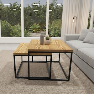 May include: Two wooden nesting coffee tables with a black metal frame. The tables have a natural wood grain finish and are placed on a beige rug. A gray sofa and large windows with a view of greenery are in the background.