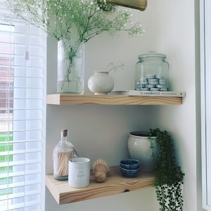 Peut inclure: Deux étagères en bois présentent des objets de décoration. Un grand vase en verre contient des fleurs blanches. On trouve également un petit vase blanc, un bocal en verre, un livre avec le mot "SCANDUSTIC" et une bougie.