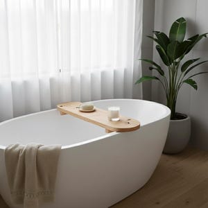 Può includere: Un vassoio da vasca in legno chiaro appoggiato su una vasca da bagno ovale bianca indipendente. Il vassoio contiene una piccola spugna rotonda beige su un piattino e una candela in un bicchiere. Un asciugamano beige pende dal bordo.