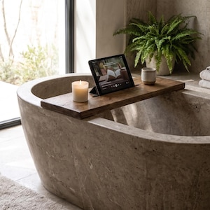 Può includere: Un vassoio rettangolare per vasca da bagno in legno marrone scuro appoggiato sopra una vasca in pietra. Il vassoio sostiene una candela bianca accesa, un tablet nero che mostra un'interfaccia digitale e una piccola tazza in ceramica.