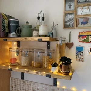 Könnte beinhalten: Zwei Holzregale mit schwarzen Metallhalterungen tragen einen NutriBullet-Mixer, eine grüne Teekanne, Vorratsgläser, Ölspender, eine Kerze und kleine Topfpflanzen. Lichterketten sind um das untere Regal gewickelt.