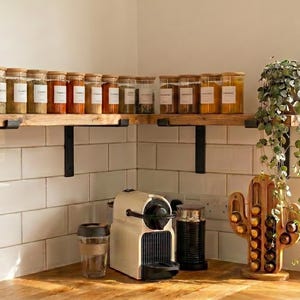 Op de afbeelding: Een houten hoekplank gemonteerd met zwarte metalen beugels, met een rij doorzichtige glazen kruidenpotjes met bamboe deksels. Daaronder staan een wit koffiezetapparaat, een glazen beker, een zwarte koffiebus en een houten cactusvormige koffiecup-houder.