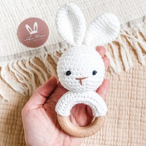 May include: A white crochet bunny rattle with a wooden ring base. The bunny has long ears, embroidered eyes, and a nose. The rattle is held against a cream-colored fabric background. The logo "Aba Mina Collection" is visible.