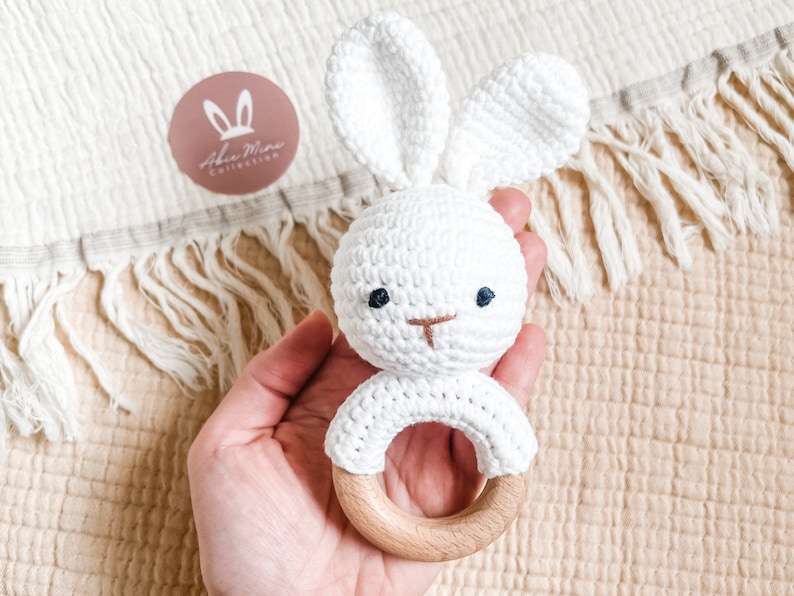 May include: A white crochet bunny rattle with a wooden ring base. The bunny has long ears, embroidered eyes, and a nose. The rattle is held against a cream-colored fabric background. The logo "Aba Mina Collection" is visible.
