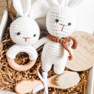 May include: A white crochet bunny rattle and a bunny plush toy with a brown scarf are in a gift box with a wooden brush and a wooden disc that says "hello world". The box is filled with brown paper.
