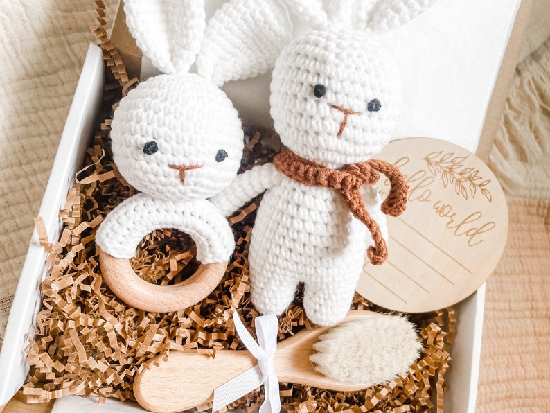 May include: A white crochet bunny rattle and a bunny plush toy with a brown scarf are in a gift box with a wooden brush and a wooden disc that says "hello world". The box is filled with brown paper.