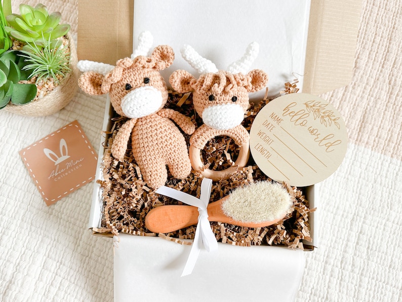 Peut inclure: Une bo&icirc;te-cadeau remplie d'articles pour b&eacute;b&eacute;, dont deux jouets en crochet en forme de vache des Highlands brun et blanc, un anneau de dentition en bois, une brosse en bois avec des poils naturels et une pancarte en bois qui dit "hello world" avec de l'espace pour le nom, la date, le poids et la taille du b&eacute;b&eacute;.