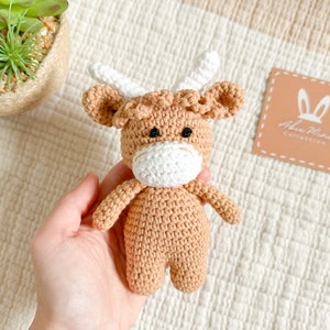 Peut inclure: Un jouet en crochet en forme de vache &eacute;cossaise, de couleur marron et blanche, avec des yeux noirs. Le jouet est tenu dans une main sur un fond textur&eacute; blanc et beige. Le fond comprend &eacute;galement une petite &eacute;tiquette carr&eacute;e avec le texte "Alia Mae Collection".