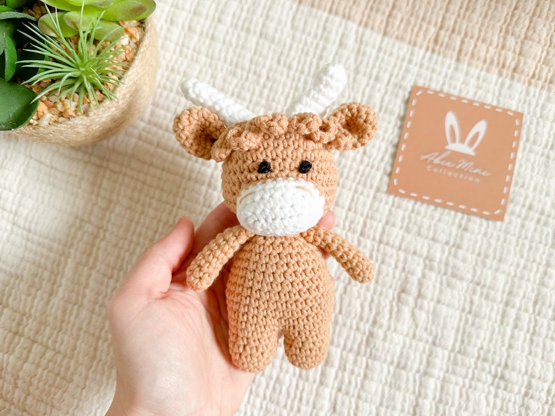 Peut inclure: Un jouet en crochet en forme de vache &eacute;cossaise, de couleur marron et blanche, avec des yeux noirs. Le jouet est tenu dans une main sur un fond textur&eacute; blanc et beige. Le fond comprend &eacute;galement une petite &eacute;tiquette carr&eacute;e avec le texte "Alia Mae Collection".