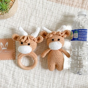 Peut inclure: Deux peluches de vache en crochet marron et blanc. Une peluche est attach&eacute;e &agrave; un anneau de dentition en bois. Les peluches sont sur une surface textur&eacute;e blanche.