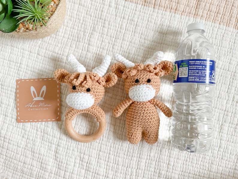 Peut inclure: Deux peluches de vache en crochet marron et blanc. Une peluche est attach&eacute;e &agrave; un anneau de dentition en bois. Les peluches sont sur une surface textur&eacute;e blanche.