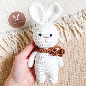May include: A handmade white crochet bunny with a brown scarf. The bunny has black eyes and a brown nose. The image includes a round logo with the text "Aben Mini Collection" and bunny ears.