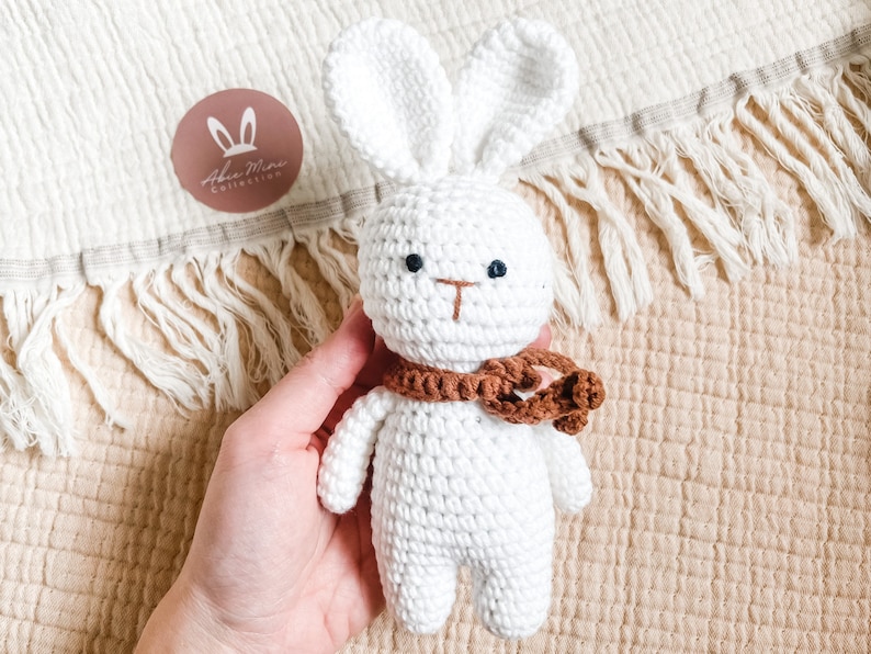 May include: A handmade white crochet bunny with a brown scarf. The bunny has black eyes and a brown nose. The image includes a round logo with the text "Aben Mini Collection" and bunny ears.