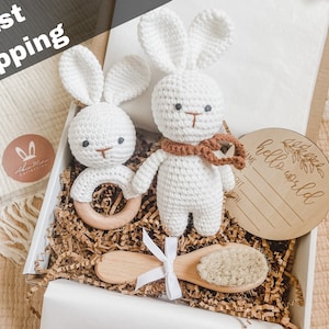 May include: A white crochet bunny rattle and a crochet bunny toy with a brown scarf are in a gift box. A wooden baby brush and a wooden sign that says "hello world" are also in the box. The words "Fast Shipping" are at the top.