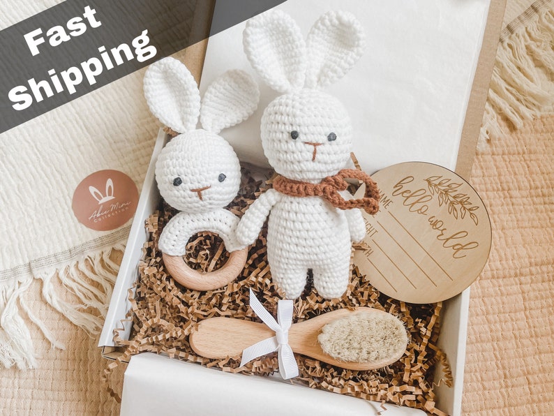 May include: A white crochet bunny rattle and a crochet bunny toy with a brown scarf are in a gift box. A wooden baby brush and a wooden sign that says "hello world" are also in the box. The words "Fast Shipping" are at the top.