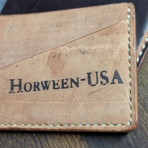 Op de afbeelding: Een lichtbruine leren kaarthouder met de tekst "HORWEEN-USA" in zwart gedrukt. De kaarthouder heeft een diagonaal designelement en is gestikt met witte draad. Andere lederwaren zijn zichtbaar op de achtergrond.