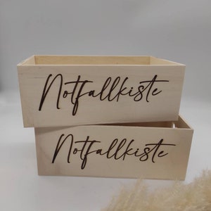May include: Two wooden boxes stacked on top of each other. Both boxes are light brown and have the words "Notfallkiste" engraved on the front in a dark brown color.