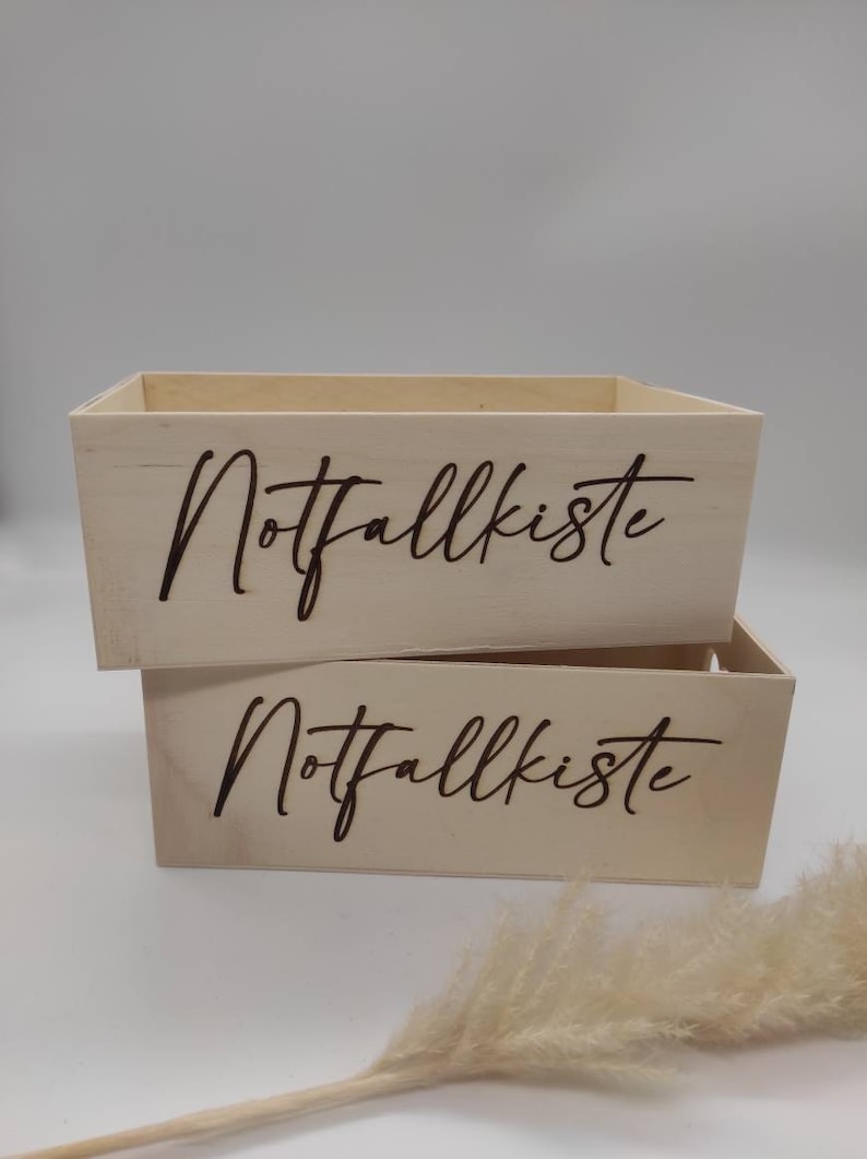 May include: Two wooden boxes stacked on top of each other. Both boxes are light brown and have the words "Notfallkiste" engraved on the front in a dark brown color.