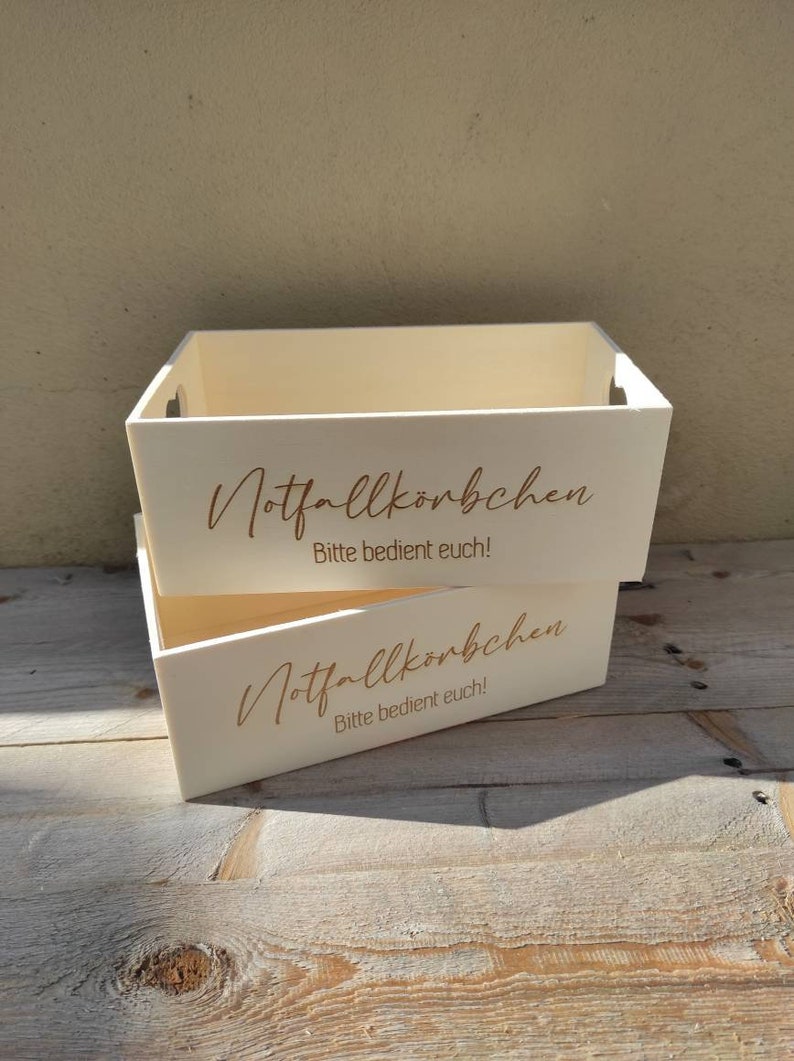 May include: Two white wooden boxes stacked on top of each other. The boxes have the text "Notfallk"rbchen" and "Bitte bedient euch!" printed on them in gold lettering.