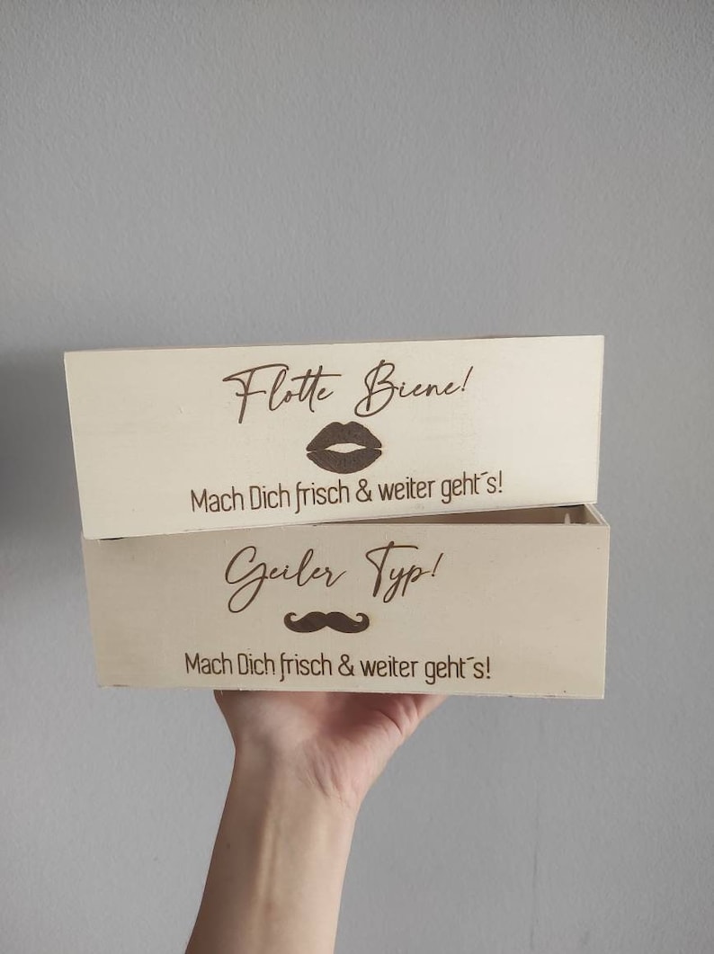 May include: Two wooden boxes with engraved messages. The top box says "Flotte Biene!" with a lipstick kiss graphic and "Mach Dich frisch & weiter geht's!" The bottom box says "Geiler Typ!" with a mustache graphic and "Mach Dich frisch & weiter geht's!"