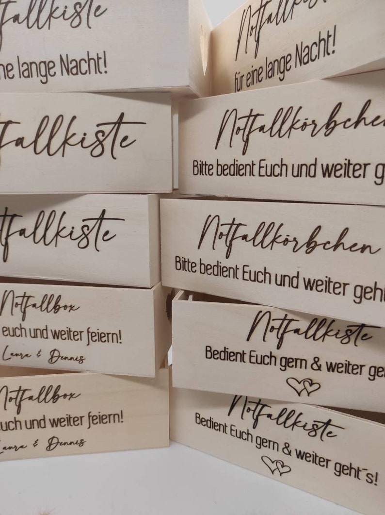May include: A stack of wooden boxes with the text "Notfallkiste" and "Notfallbox" engraved on them. The boxes are decorated with hearts and the text "Bedient Euch gern & weiter geht's!"