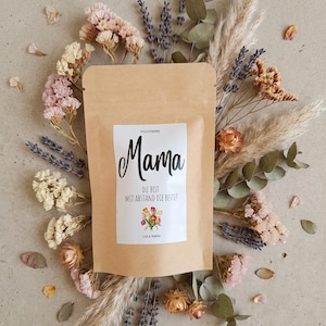 Könnte beinhalten: Ein brauner Papierteebeutel mit dem Wort "Mama" in schwarzer Schrift, umgeben von getrockneten Blumen in Rosa-, Creme- und Lavendeltönen. Der Beutel hat ein weißes Etikett mit deutschem Text und einer floralen Illustration. Die Anordnung befindet sich vor einem neutralen Hintergrund.