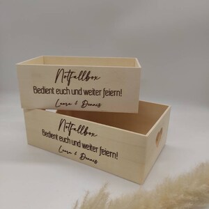 May include: Two wooden boxes with a heart-shaped cutout on the side. The boxes are labeled "Notfallbox" and have the text "Bedient euch und weiter feiern!" and "Laura & Dennis" engraved on them.