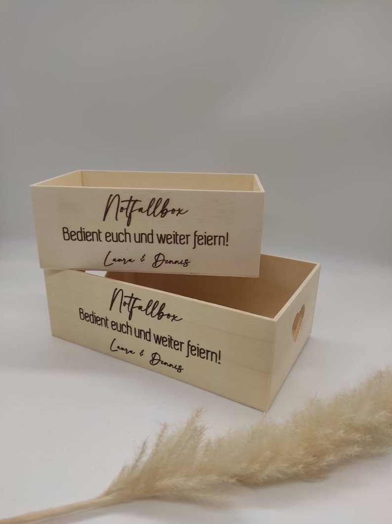May include: Two wooden boxes with a heart-shaped cutout on the side. The boxes are labeled "Notfallbox" and have the text "Bedient euch und weiter feiern!" and "Laura & Dennis" engraved on them.