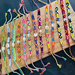 May include: A collection of twelve handmade woven friendship bracelets featuring intricate geometric patterns in vibrant colors including pink, yellow, blue, purple, and brown. Each bracelet is accented with a single white cowrie shell at the center.
