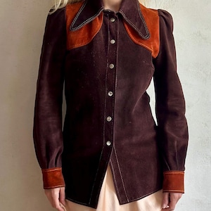 Puede incluir: Una camisa de ante marrón oscuro con un canesú y puños de ante naranja en contraste. La camisa tiene cuello puntiagudo, mangas largas y cierre de botones en la parte delantera. Las costuras son de un color más claro.
