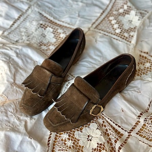 May include: A pair of brown suede loafers with fringe detailing and gold-colored buckles. The shoes have a classic design and are set against a white, patterned background.