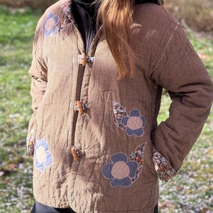 May include: Brown quilted jacket with a small checkered pattern, decorated with blue floral appliqués. The jacket has wooden toggle closures and patterned cuffs, offering a unique, handcrafted aesthetic. A stylish outerwear piece.