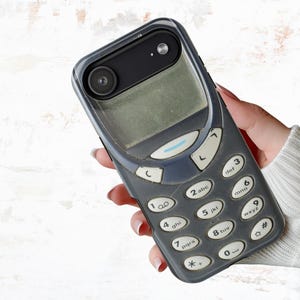 May include: A phone case with a retro design resembling an old cell phone. The case is dark gray with a keypad and screen, and has a black camera lens. The case is held in a hand.
