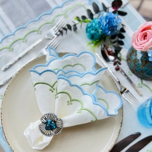 Peut inclure: Une serviette en lin blanc avec une bordure brodée bleue et verte est pliée et maintenue ensemble par un anneau de serviette en argent avec un bijou bleu. La serviette est sur une assiette blanche avec un set de table brodé bleu et vert.