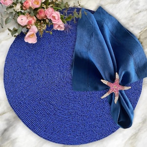 May include: A blue beaded placemat with a blue napkin and a pink jeweled starfish napkin ring.