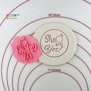 Può includere: Due stampini per biscotti in silicone rosa. Uno stampino ha la forma di un anello con le parole "She Said Yes!" e l'altro stampino ha la forma di un anello con le parole "She Said Yes!" e un diamante.