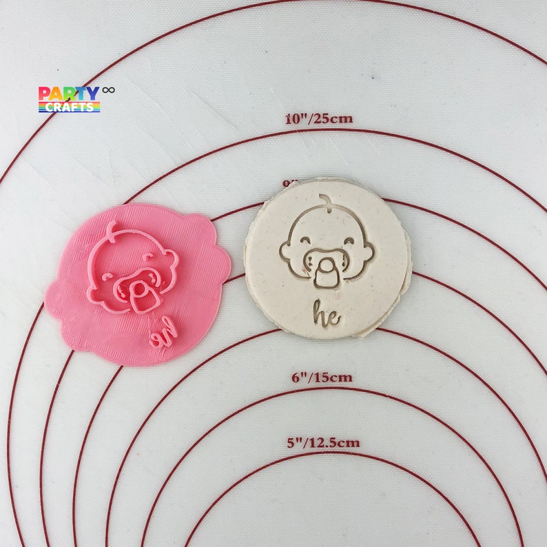 May include: A pink and white baby cookie cutter with a baby face and pacifier design. The cutter is 6 inches in diameter and is shown on a white silicone baking mat with red concentric circles.