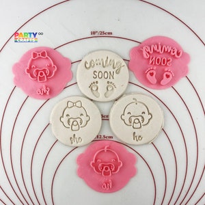 Può includere: Set di sei tagliabiscotti a tema bambino in rosa e bianco. I tagliabiscotti hanno la forma di un viso di bambino, un piede di bambino e uno striscione che dice "coming soon".