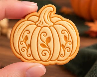 May include: A light tan, pumpkin-shaped cookie featuring a raised, embossed design of swirling vines and leaves on its surface. The cookie has a smooth, baked texture with defined ridges and a prominent stem at the top, showcasing intricate details.