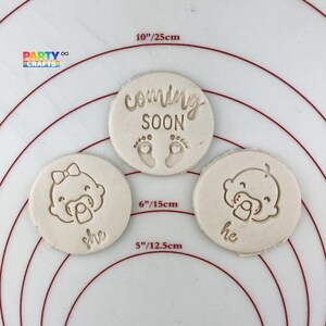 May include: Three round, white, clay cookie cutters. The largest cutter is 10 inches in diameter and reads "coming soon" with two small footprints. The two smaller cutters are 6 inches and 5 inches in diameter. The smaller cutters have a baby face with a pacifier and the words "she" and "he" underneath.
