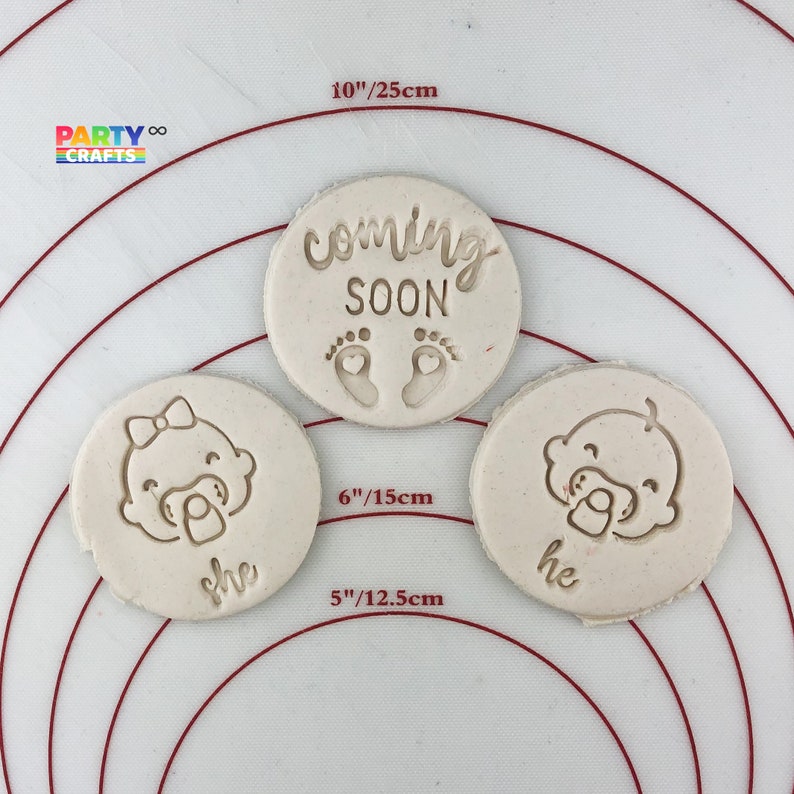 May include: Three round, white, clay cookie cutters. The largest cutter is 10 inches in diameter and reads "coming soon" with two small footprints. The two smaller cutters are 6 inches and 5 inches in diameter. The smaller cutters have a baby face with a pacifier and the words "she" and "he" underneath.